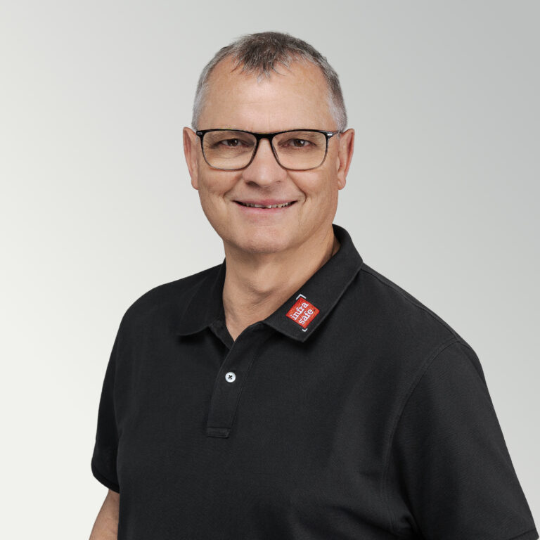 Ein lächelnder Mann mittleren Alters mit kurzen grauen Haaren und Brille trägt ein schwarzes Poloshirt mit einem roten Namensschild und steht vor einem hellgrauen Hintergrund mit Farbverlauf.