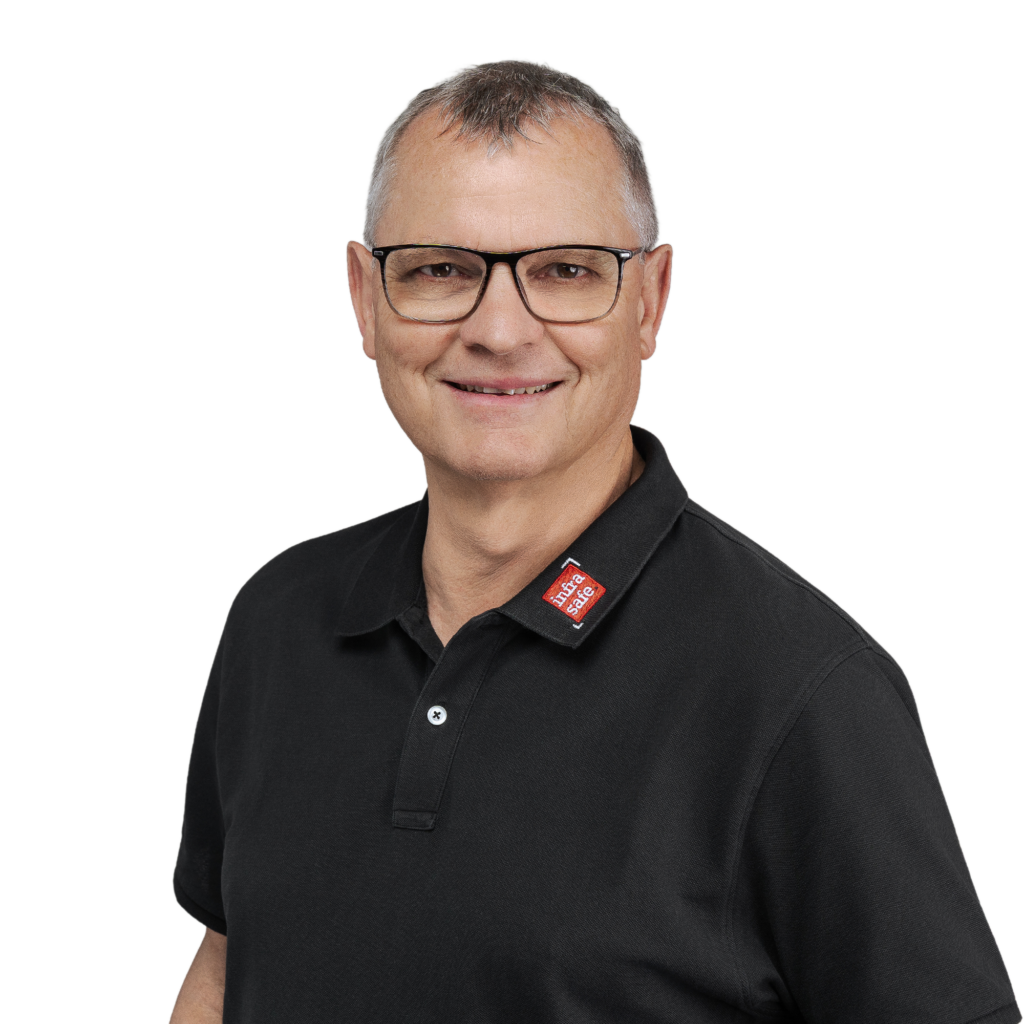 Ein lächelnder Mann mit kurzen grauen Haaren und Brille, der ein schwarzes Poloshirt und ein rotes Namensschild trägt und vor einem schlichten weißen Hintergrund steht.