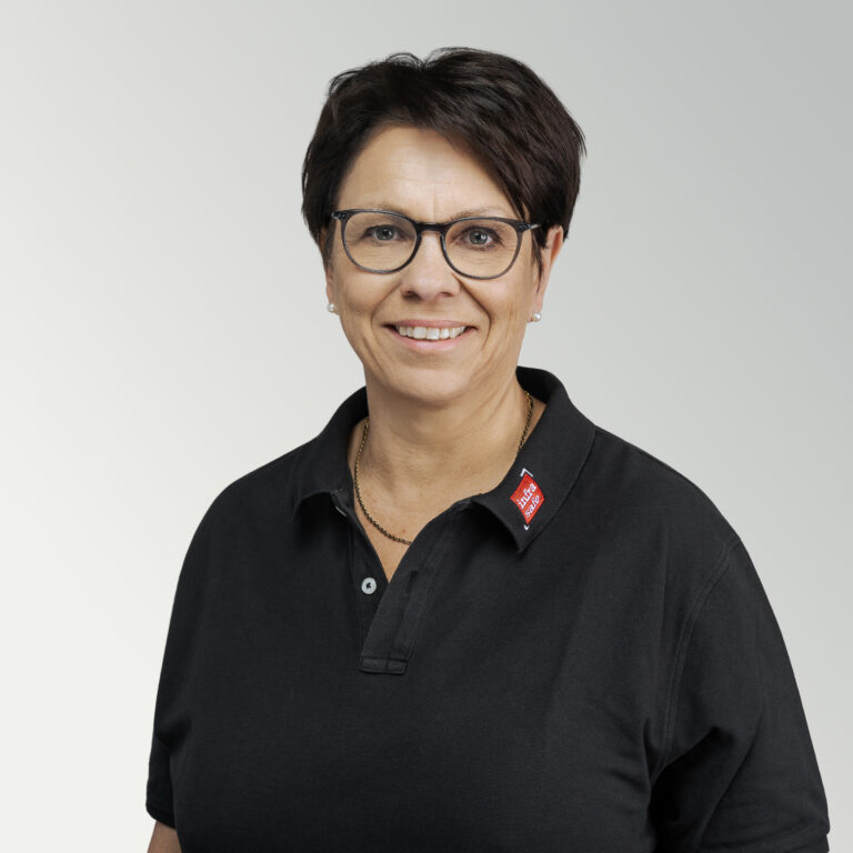 Eine Frau mit kurzen dunklen Haaren, die eine Brille trägt und ein schwarzes Hemd mit Kragen und einem roten Namensschild, lächelt vor einem hellgrauen Hintergrund mit Farbverlauf in die Kamera.