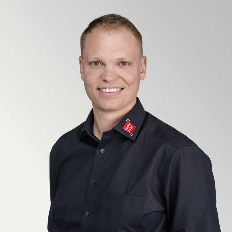 Eine lächelnde Person mit kurzen blonden Haaren, die ein schwarzes Hemd mit Kragen und einem rot-weißen Logo am Kragen trägt und vor einem hellgrauen Hintergrund steht.